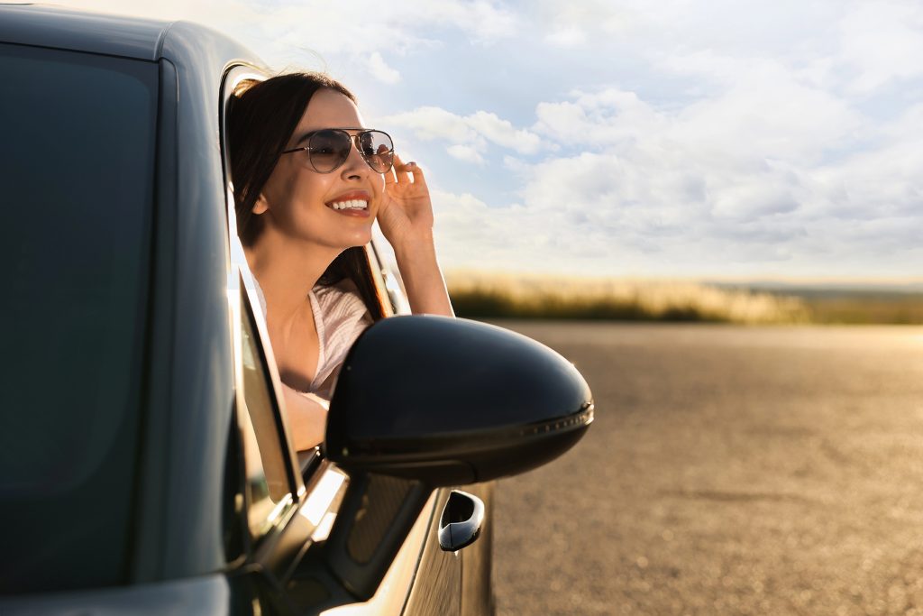 smiling young woman in sunglasses leaning out of car window outd